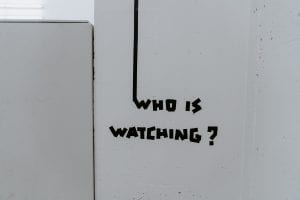 White wall with “Who Is Watching?” written in black electrical tape; image by Claudio Schwarz, via Unsplash.com.